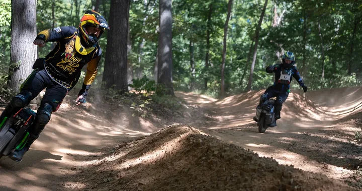 Two electric unicycle racers corner on a wooded dirt course at Camp Shred.