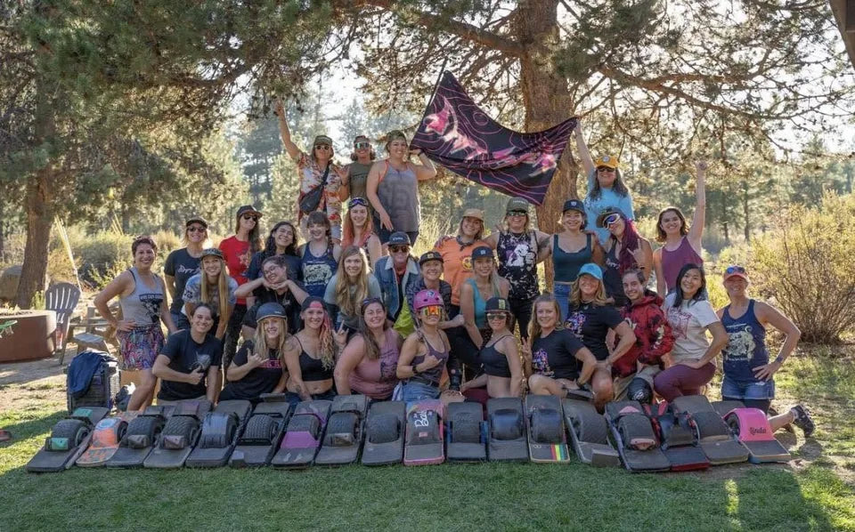 Drift Sisters riders pose behind a row of Onewheels beneath their flag.