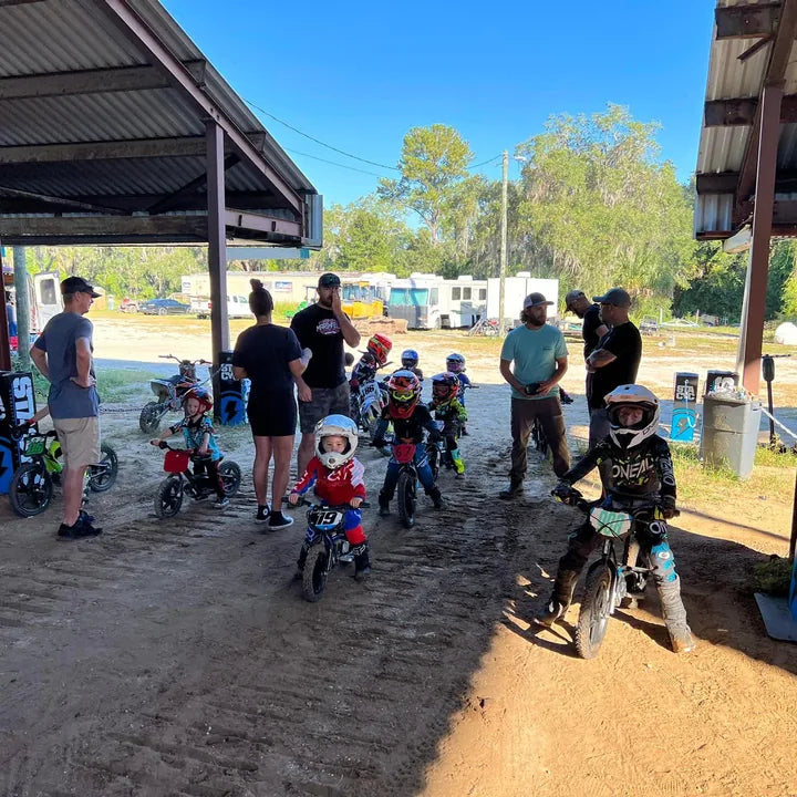Young riders on Stacycs gather with adults beneath a covered dirt-track staging area at E Jam.