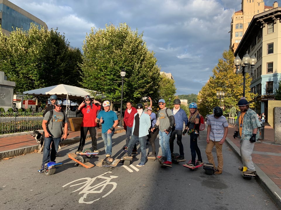 Twelve helmeted riders pose downtown with a dog and their Onewheels beneath dramatic clouds.