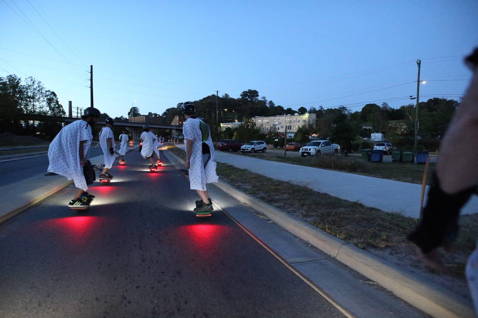 Helmeted Onewheel riders in white gowns ride together at dusk with red tail lights reflecting on the road.