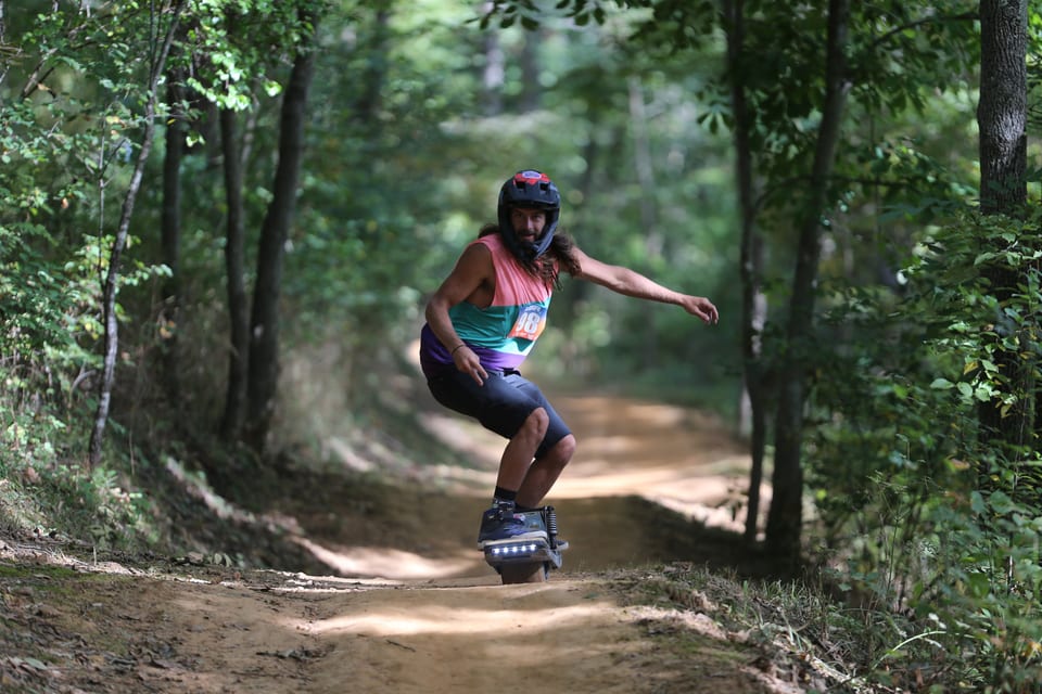 A helmeted rider wearing number 98 carves toward the camera on a wooded dirt trail.