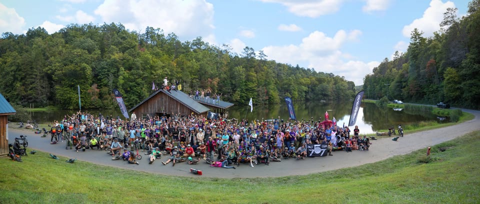 A panoramic crowd of Onewheel riders gathers on a grassy hillside beside the lake.