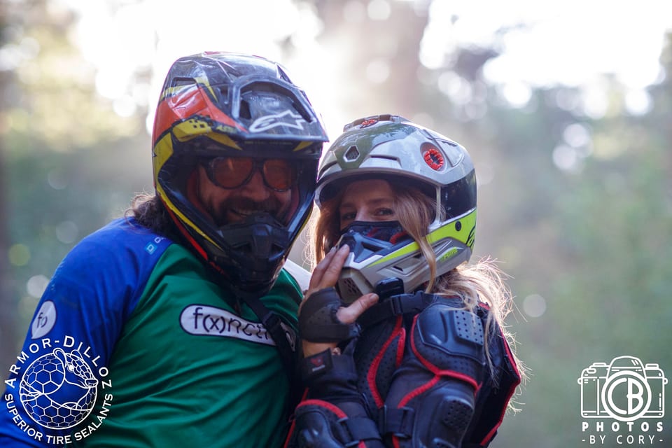 Two smiling helmeted riders stand shoulder to shoulder in the woods at Seek N' Shred.
