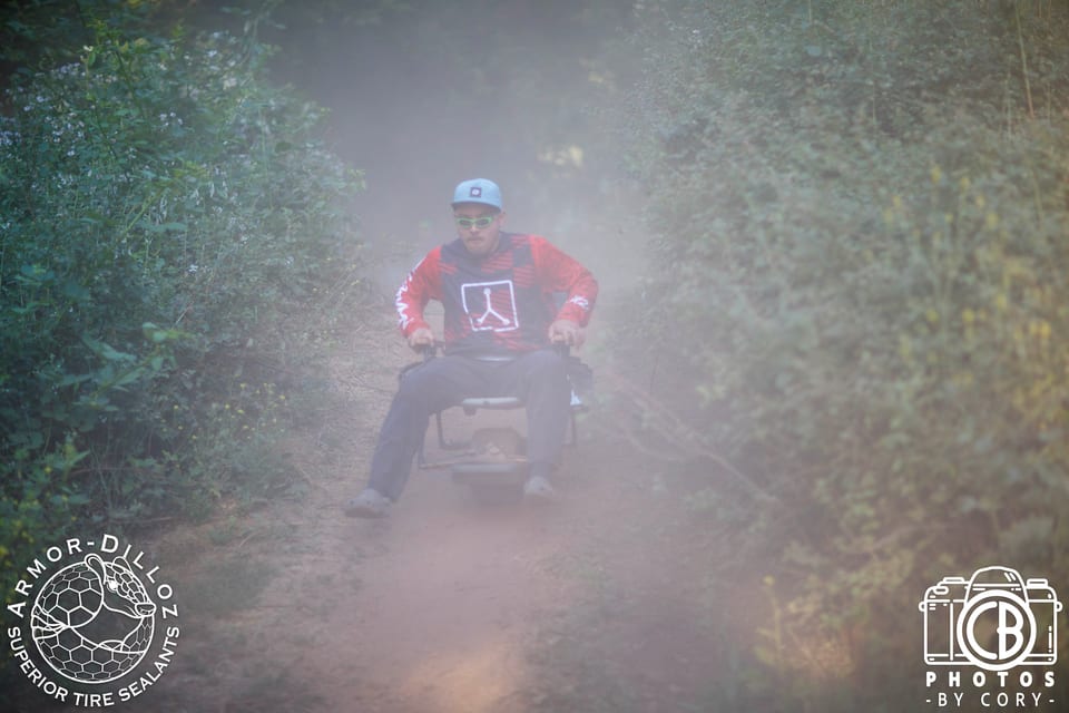 A helmeted racer rides a wheeled lawn chair through a dust cloud with both arms out.