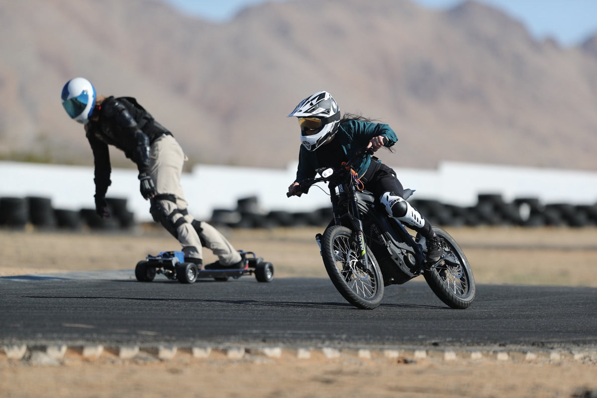 An electric bike racer and an electric skateboard racer lean through a paved turn with mountains behind them.