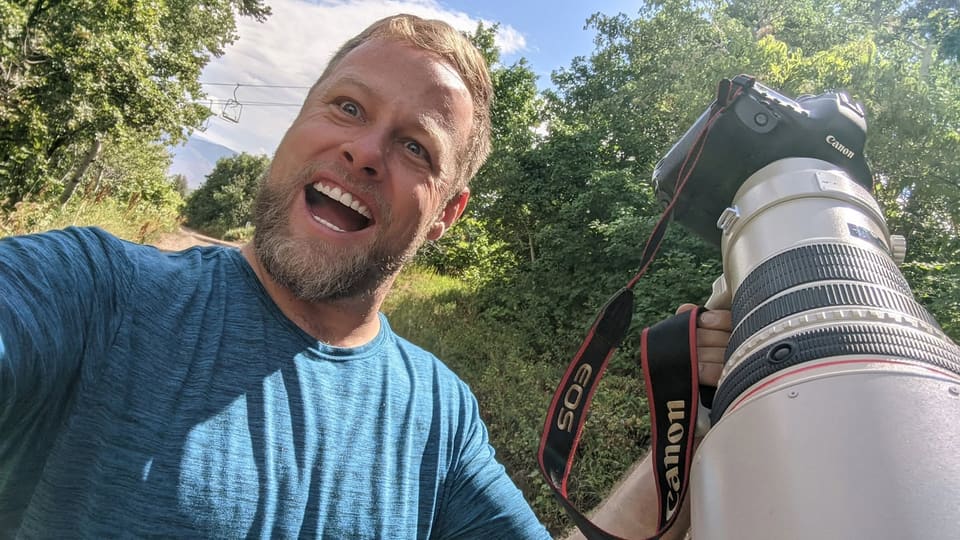 Cory grinning beside his camera on a wooded trail.