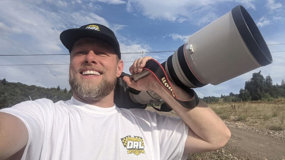 Cory smiling with a camera and long white telephoto lens resting on his shoulder.